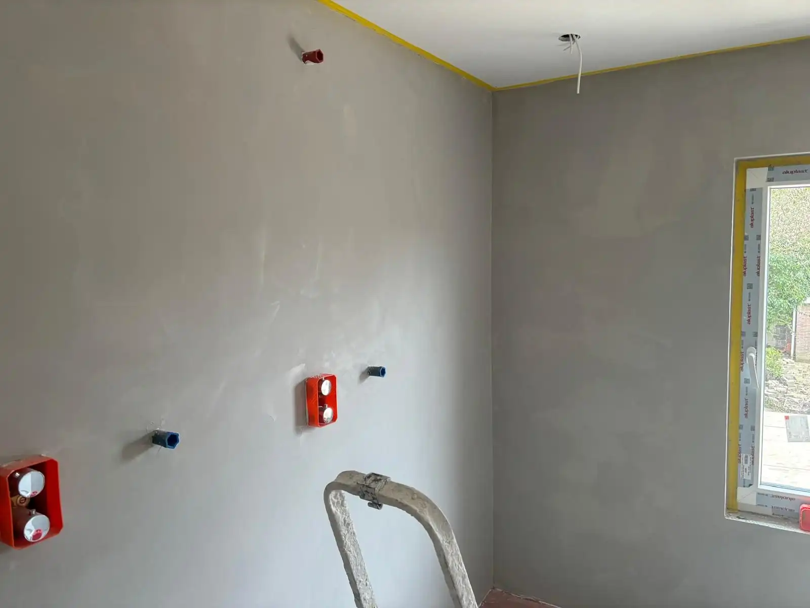 Vers gestucte grijze wand naast een raam, met oranje inbouwdozen en afgeplakte plafondrand.