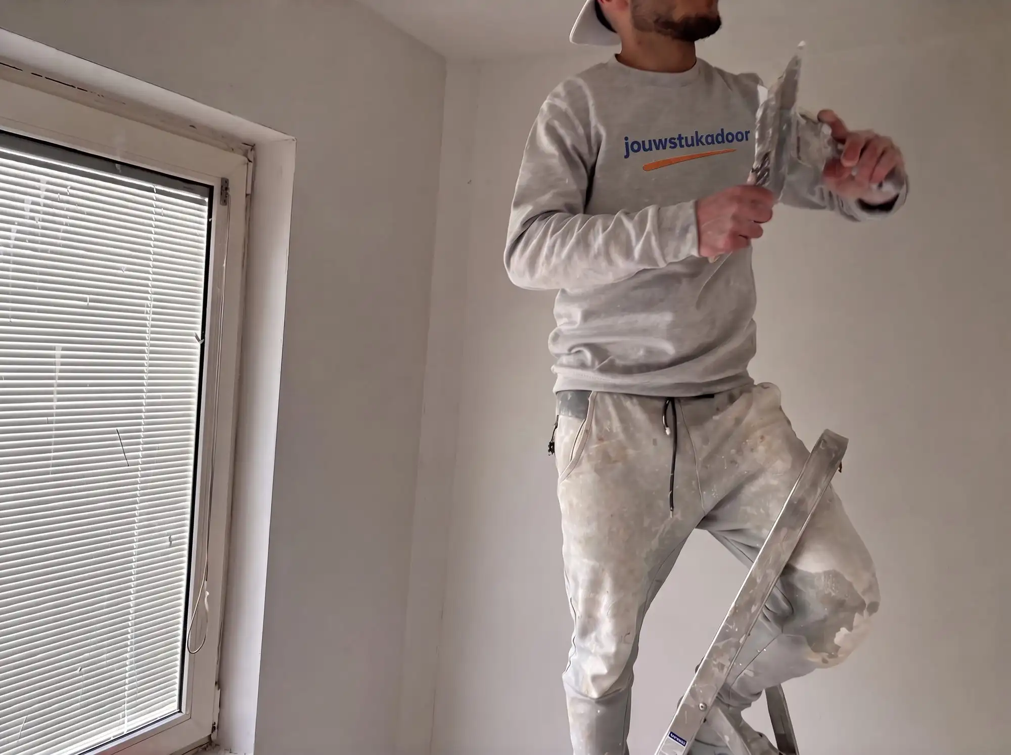 Stukadoor van jouwstukadoor op een trapje met spaan en plakspaan, naast een raam.
