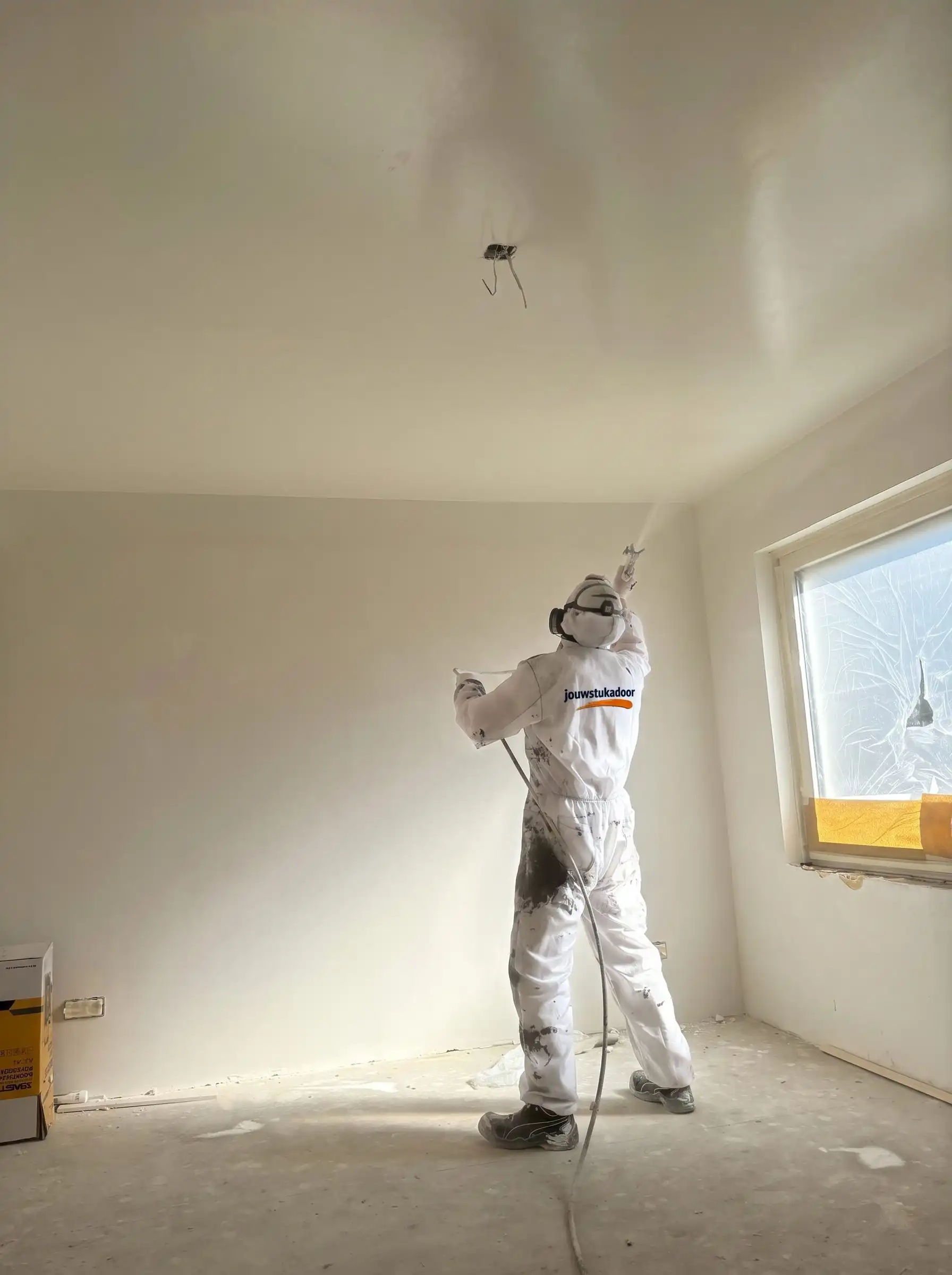 Stukadoor van jouwstukadoor in witte overall spuit een plafond af in een lichte kamer.